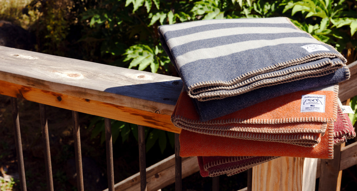 All three Northwest Wool Throw colors (Denim Blue, Mesa Sunset, and Rainier Red) folded and stacked on a back deck.