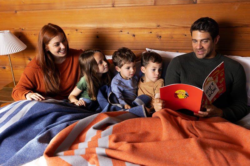 Family snuggled in bed with their Denim Blue and Mesa Sunset Northwest Wool Throws reading a book together.